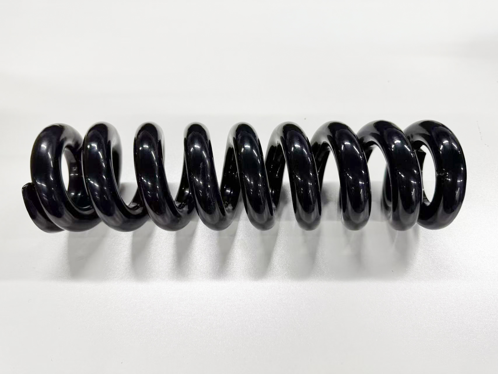 Black helical compression spring resting horizontally on a white surface.